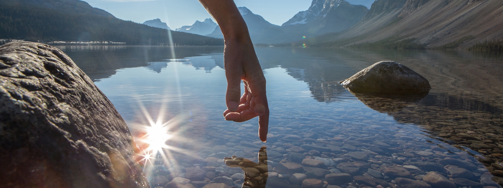 Finger touches surface of mountain lake. The sun and the landscape is reflecting on the water.